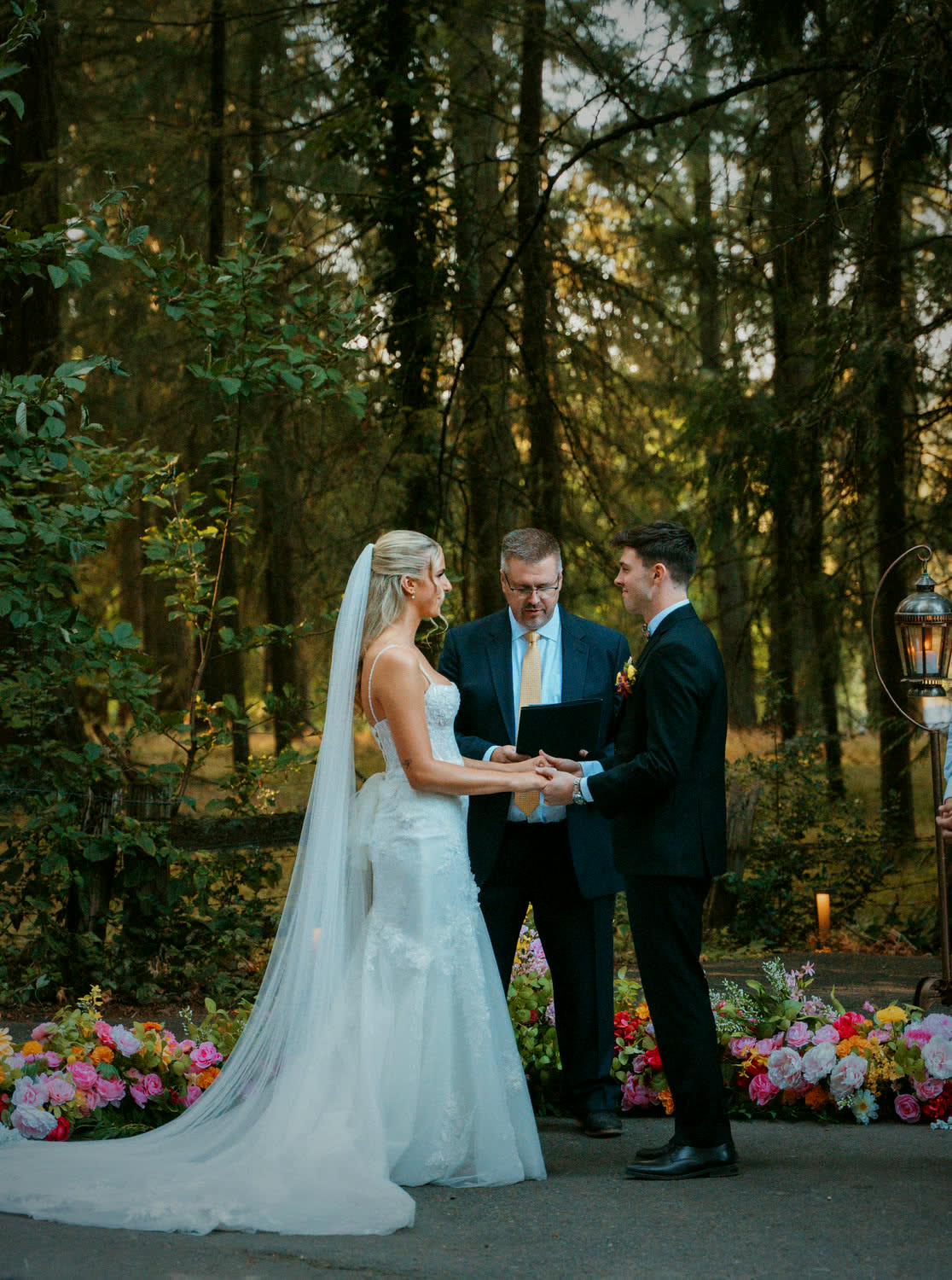 A couple exchanging vows on the forest aisle, banked flowers along the front row