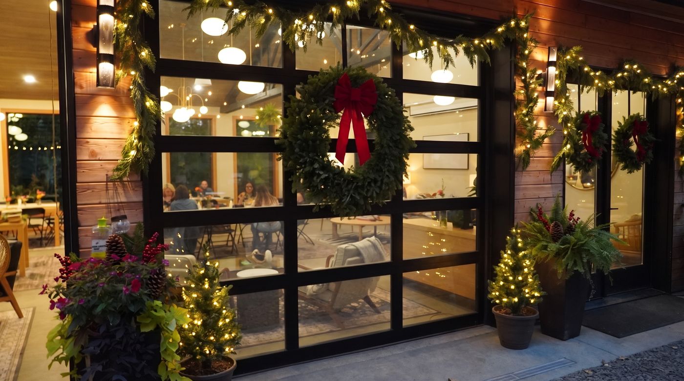 The hall's glass garage door and entry door at night, each hung with an evergreen wreath and a red bow, garland and small lit trees beside them