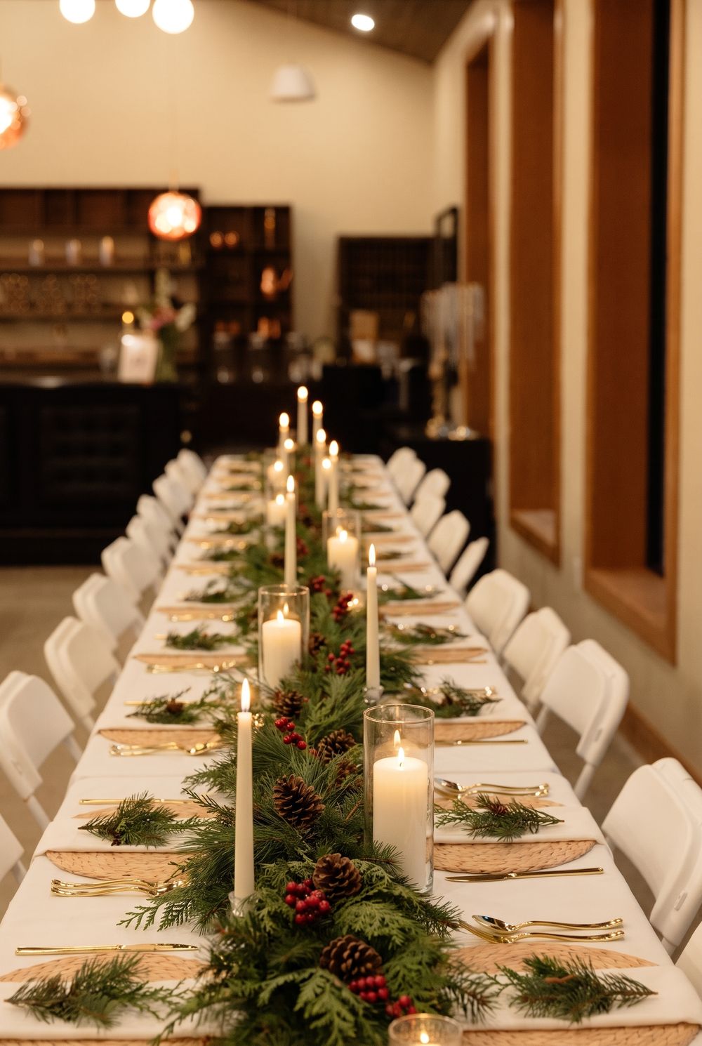A long dinner table down the hall with pine and cedar garland, pinecones, red berries and white candles in glass hurricanes